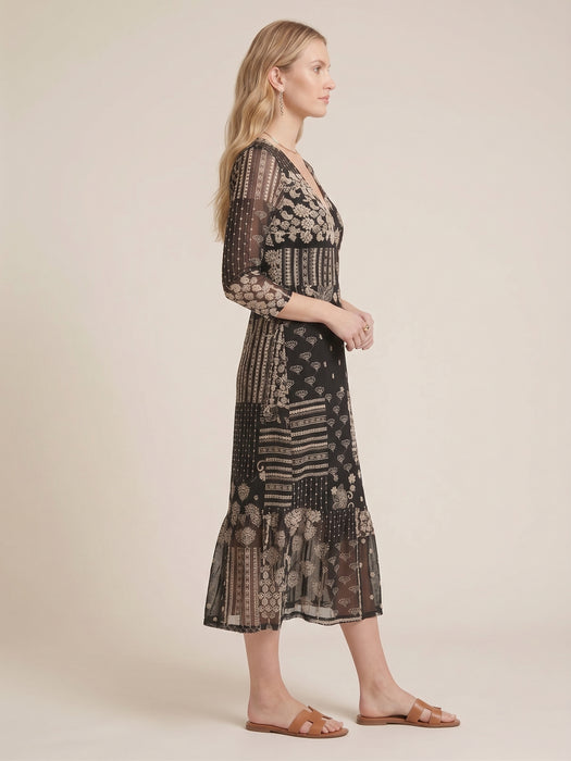 A woman modeling a long-sleeved midi dress with a black and beige patchwork-style bohemian print, shown in profile against a solid beige studio background.
