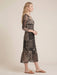 A woman modeling a long-sleeved midi dress with a black and beige patchwork-style bohemian print, shown in profile against a solid beige studio background.