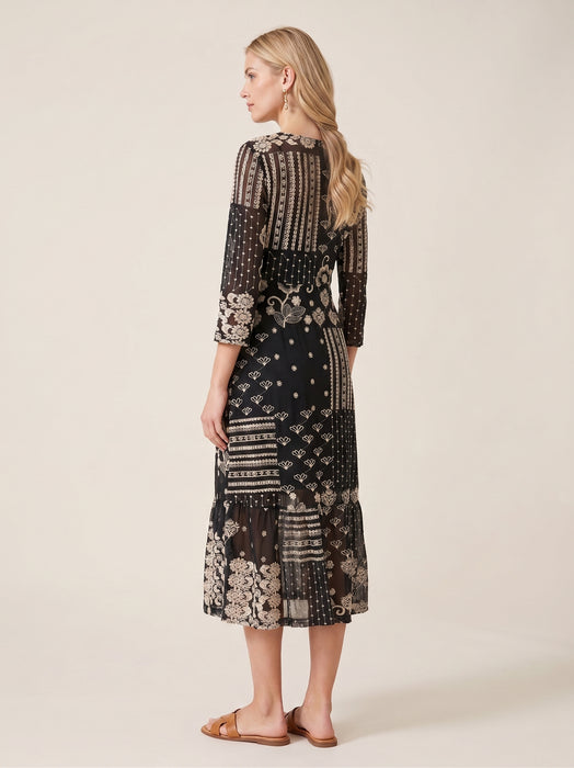 A woman modeling a black midi dress with beige and white patchwork-style prints, shown from a side-back angle against a plain beige studio background.