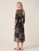 A woman modeling a black midi dress with beige and white patchwork-style prints, shown from a side-back angle against a plain beige studio background.