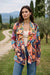 A woman stands outdoors in a grassy field with a dirt path and trees in the background, wearing a colorful bohemian floral print sherpa jacket with slip pockets from Johnny Was