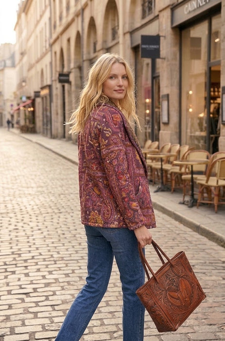 A woman walking on a cobblestone street wearing a rust-brown paisley patterned jacket and blue jeans, carrying a brown leather tote bag.