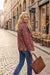 A woman walking on a cobblestone street wearing a rust-brown paisley patterned jacket and blue jeans, carrying a brown leather tote bag.