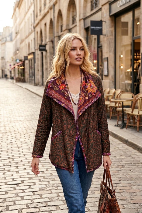 A woman walking down a cobblestone street wearing a brown patterned jacket with colorful embroidered details on the collar, blue jeans, and carrying a brown leather handbag.