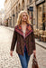 A woman walking down a cobblestone street wearing a brown patterned jacket with colorful embroidered details on the collar, blue jeans, and carrying a brown leather handbag.