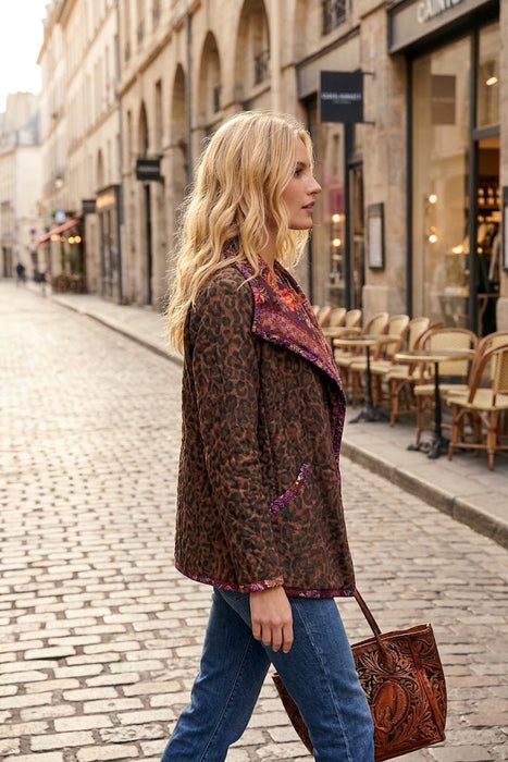 A woman walking on a cobblestone street wearing a brown leopard-print jacket with a purple and red patterned lining, blue jeans, and carrying a brown leather handbag.