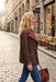 A woman walking on a cobblestone street wearing a brown leopard-print jacket with a purple and red patterned lining, blue jeans, and carrying a brown leather handbag.