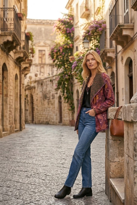 A woman stands on a cobblestone street in a historic town, wearing a patterned burgundy and brown jacket, blue jeans, black ankle boots, and leaning against a stone wall with a brown shoulder bag.