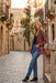 A woman stands on a cobblestone street in a historic town, wearing a patterned burgundy and brown jacket, blue jeans, black ankle boots, and leaning against a stone wall with a brown shoulder bag.