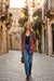 A woman walking down a cobblestone street in an old European town, wearing a patterned bohemian-style jacket with a brown suede-like collar, blue jeans, and black ankle boots.