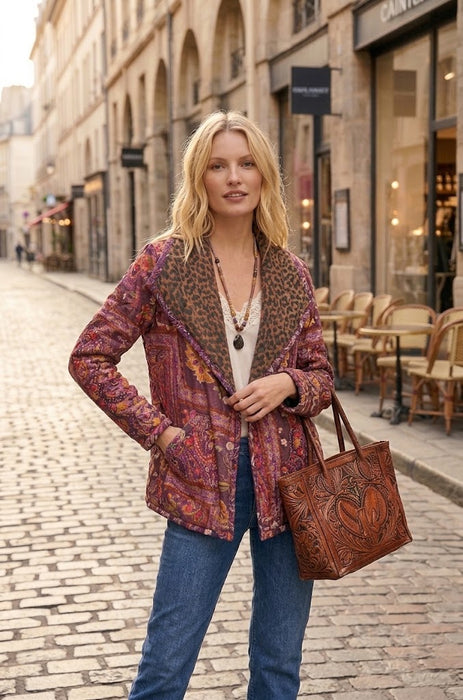 A woman stands on a cobblestone street wearing a patterned brown and red jacket with a leopard-print collar, blue jeans, and carrying a large brown tooled leather tote bag.