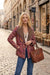 A woman stands on a cobblestone street wearing a patterned brown and red jacket with a leopard-print collar, blue jeans, and carrying a large brown tooled leather tote bag.