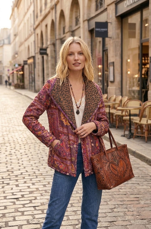 A woman stands on a cobblestone street wearing a patterned brown and red jacket with a leopard-print collar, blue jeans, and carrying a large brown tooled leather tote bag.