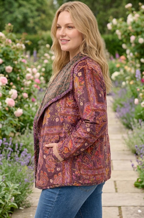A woman wearing a long-sleeve quilted jacket with a multicolor paisley pattern in purple, orange, and brown, standing in a garden pathway.