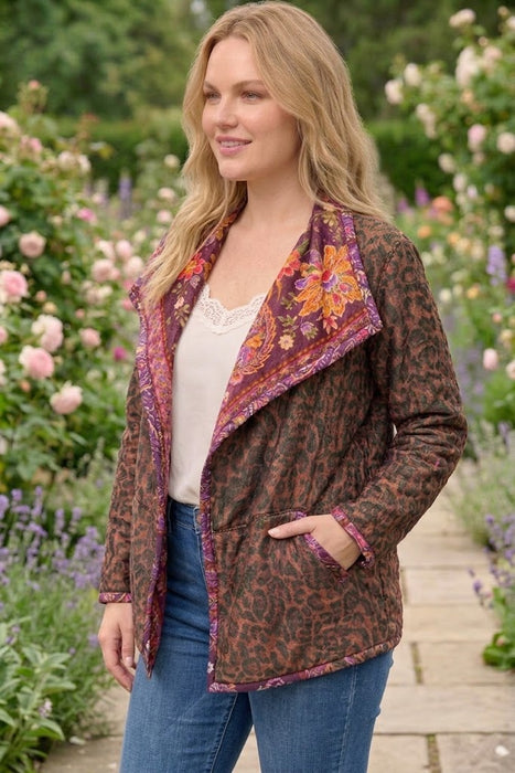 A woman wearing a brown leopard-print jacket with purple and orange paisley trim, styled with a white top and blue jeans, standing in a garden path.