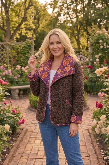 A woman stands in a garden wearing a brown leopard-print jacket with purple and orange patterned lining, blue jeans, and a white top. The background shows a brick pathway and lush green foliage with pink and white flowers.