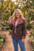 A woman stands in a garden wearing a brown leopard-print jacket with purple and orange patterned lining, blue jeans, and a white top. The background shows a brick pathway and lush green foliage with pink and white flowers.