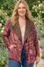 A woman standing in a garden wearing a patterned open-front jacket with a leopard-print collar, layered over a cream top and blue jeans.