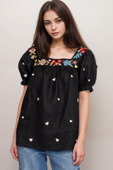 Woman wearing a black blouse with colorful floral and heart embroidery, square neck, and puff sleeves