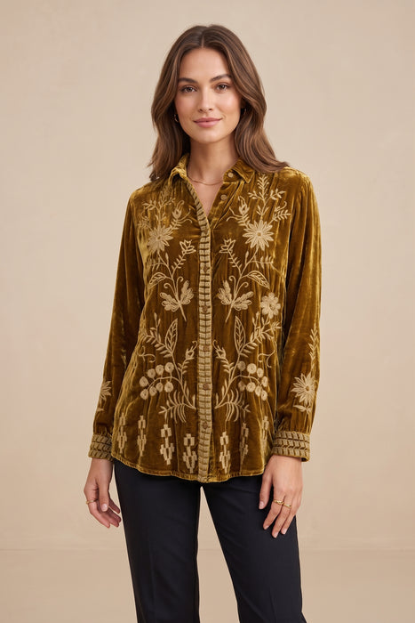 A woman modeling a long-sleeve button-up shirt made of rich olive-brown velvet with intricate cream floral embroidery, paired with black pants, against a solid beige studio background.