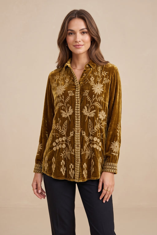 A woman modeling a long-sleeve button-up shirt made of rich olive-brown velvet with intricate cream floral embroidery, paired with black pants, against a solid beige studio background.