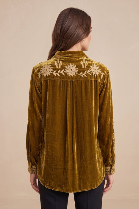 A woman viewed from the back wearing a mustard yellow velvet long-sleeve shirt with white floral embroidery across the shoulders, paired with black pants, against a solid beige background.