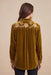A woman viewed from the back wearing a mustard yellow velvet long-sleeve shirt with white floral embroidery across the shoulders, paired with black pants, against a solid beige background.