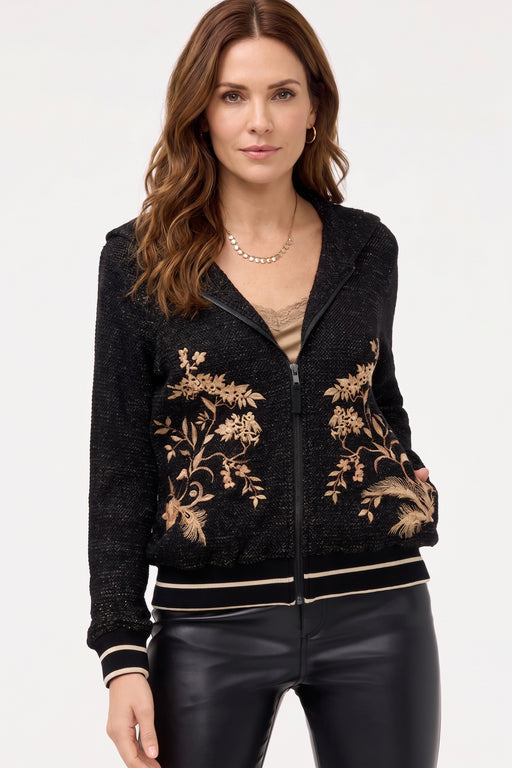 A woman wearing a black zip-up hoodie jacket with beige floral embroidery on the front, paired with black faux-leather pants, against a plain white studio background.