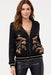 A woman wearing a black zip-up hoodie jacket with beige floral embroidery on the front, paired with black faux-leather pants, against a plain white studio background.