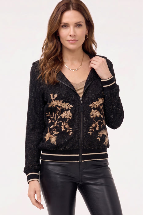 A woman modeling a black zip-up knit jacket with beige floral embroidery on the front, ribbed trim, and black leather-look pants against a plain light background.