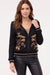 A woman modeling a black zip-up knit jacket with beige floral embroidery on the front, ribbed trim, and black leather-look pants against a plain light background.