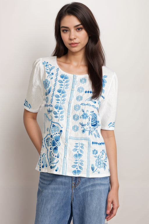 Woman wearing a white linen blouse with blue embroideries and button front from Johnny Was