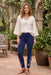 A woman stands in a garden courtyard wearing navy blue leggings with colorful floral embroidery near the ankles, a cream linen blouse, and brown sandals.