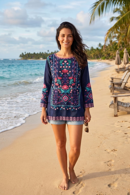 A woman walking on a sandy beach wearing a navy blue embroidered tunic top with colorful floral patterns and light blue denim shorts.