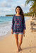 A woman walking on a sandy beach wearing a navy blue embroidered tunic top with colorful floral patterns and light blue denim shorts.