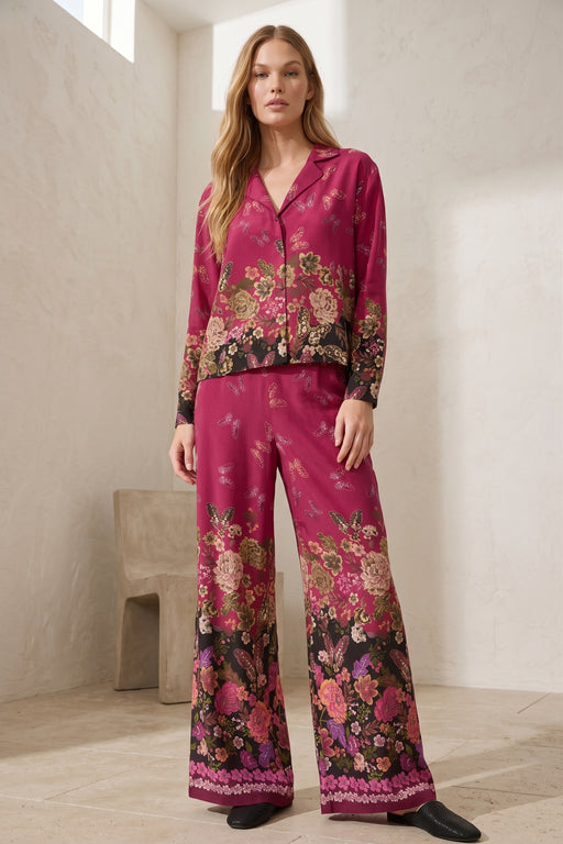 A woman modeling a two-piece pajama set with a vibrant magenta floral print and lace trim, standing in a minimalist cream-colored room.