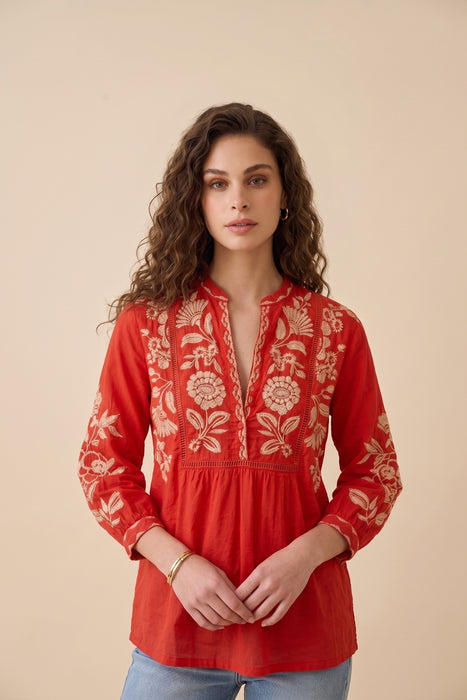 A woman wearing a red long-sleeve blouse with beige floral embroidery, paired with light blue jeans, standing against a solid beige background.