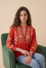 A woman with long curly brown hair sits in a green velvet armchair, wearing a vibrant red cotton blouse with intricate cream floral embroidery and light blue jeans, set against a solid beige background.