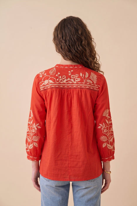 A woman viewed from the back wearing a bright red long-sleeve blouse with white floral embroidery across the yoke and sleeves, paired with light blue jeans against a solid beige background.