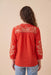 A woman viewed from the back wearing a bright red long-sleeve blouse with white floral embroidery across the yoke and sleeves, paired with light blue jeans against a solid beige background.