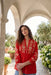 A woman wearing a vibrant red long-sleeve blouse with intricate gold floral embroidery, standing under a stone archway in a garden setting.