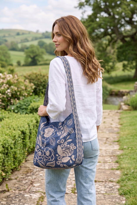 A woman wearing a white long-sleeve top and light blue jeans stands on a stone path in a garden, carrying a large navy blue shoulder bag with a beige floral pattern and a patterned strap.