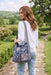 A woman wearing a white long-sleeve top and light blue jeans stands on a stone path in a garden, carrying a large navy blue shoulder bag with a beige floral pattern and a patterned strap.