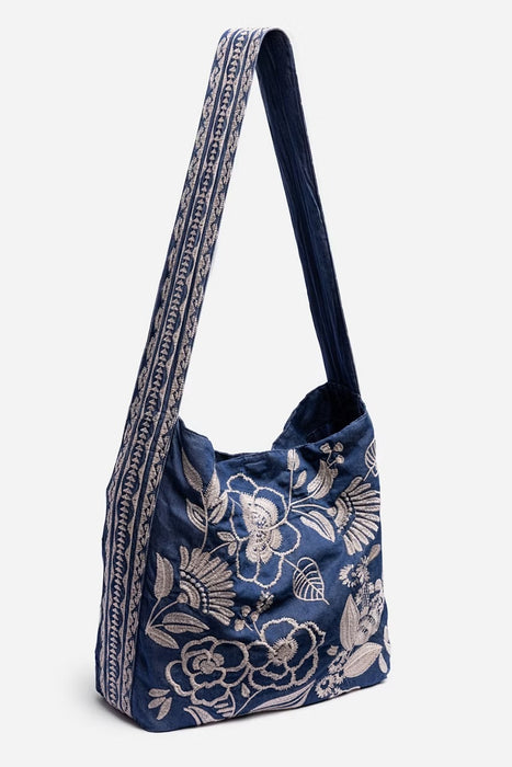 A navy blue fabric shoulder bag with a white floral and bird pattern, featuring a wide shoulder strap with a white geometric border, set against a light gray background.
