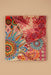 A folded multicolor paisley and floral scarf with intricate patterns in red, pink, blue, orange, and yellow, displayed on a solid beige background.
