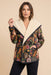 A woman models a long-sleeve jacket featuring a cream-colored sherpa collar and a dark multicolor floral and paisley patterned body, set against a solid beige background.