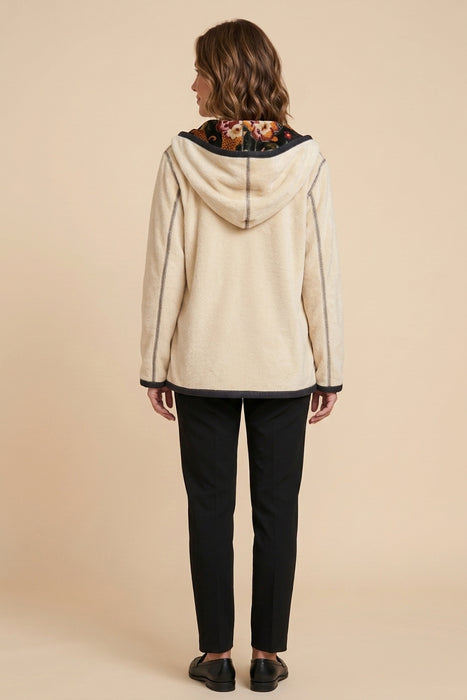A woman viewed from the back wearing a cream-colored hooded jacket with a dark floral lining and black pants, set against a solid beige studio background.