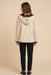 A woman viewed from the back wearing a cream-colored hooded jacket with a dark floral lining and black pants, set against a solid beige studio background.