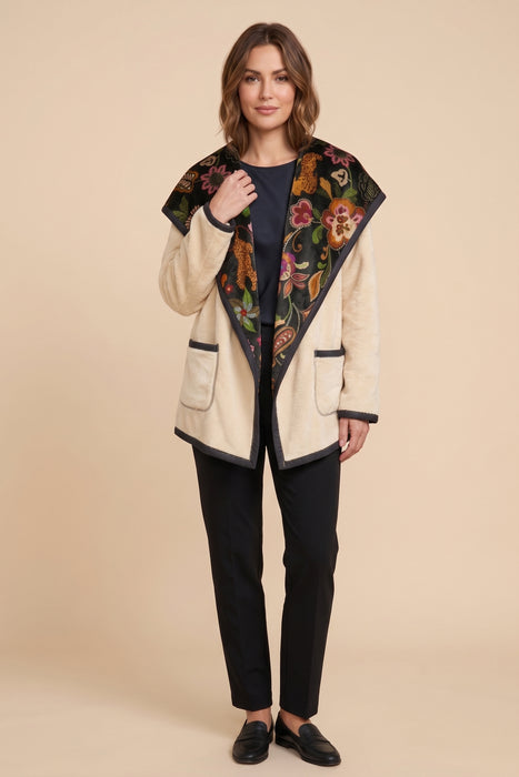 A woman modeling a cream-colored open-front jacket with a large black floral-patterned collar, paired with black trousers and black loafers, against a solid beige background.
