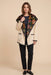 A woman modeling a cream-colored open-front jacket with a large black floral-patterned collar, paired with black trousers and black loafers, against a solid beige background.